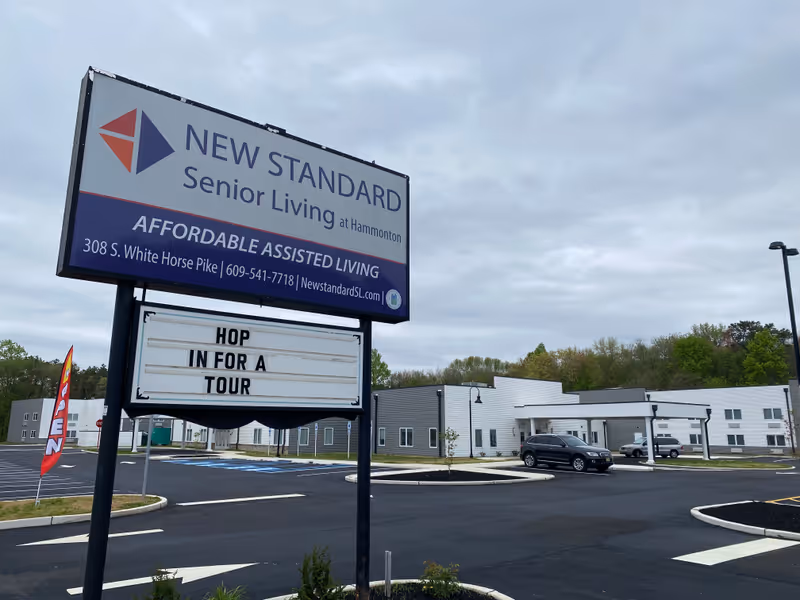 Exterior view of New Standard Senior Living at Hammonton facility with a large sign displaying the name, address, and contact information. The sign also has a message board that reads 'HOP IN FOR A TOUR'. The building is modern with white and gray walls, surrounded by a parking lot with a few cars and an 'OPEN' flag near the entrance. The sky is overcast.