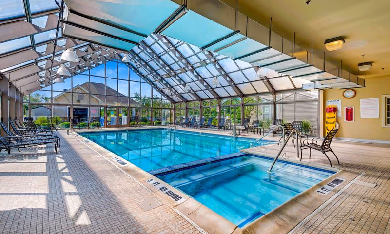 Sunlit indoor swimming pool and adjoining hot tub inside a glass-enclosed atrium with lounge chairs.