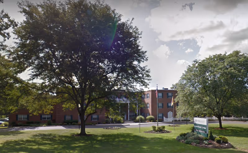 Exterior view of a brick senior living facility building with multiple windows, surrounded by large trees and a well-maintained lawn. There is a sign near the entrance that reads 'Green Knoll Center' and two flagpoles with flags in front of the building under a partly cloudy sky.