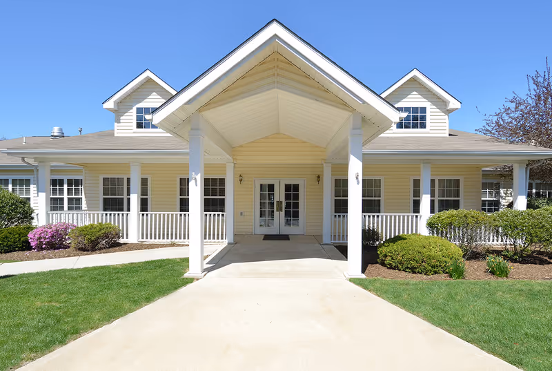 Front exterior view of a single-story building with a covered entrance supported by four white columns. The building has light yellow siding, white trim, multiple windows, and a well-maintained lawn with bushes and flowers on either side of the entrance walkway. The sky is clear and blue.