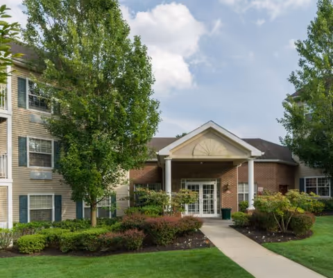 Front entrance of a residential building with a covered portico, walkway, and landscaped lawn and trees.