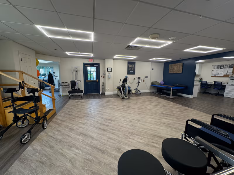 Large rehabilitation room with exercise machines, walkers, a treatment table and seating in a senior care facility.