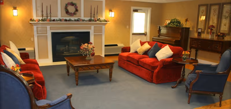 A cozy living room in an assisted senior living facility featuring two red sofas with multiple cushions, two blue armchairs, a wooden coffee table with a floral centerpiece, a fireplace decorated with a wreath and candles, a piano, and a wooden sideboard with framed artwork and flowers.