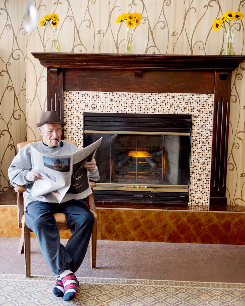 An elderly man wearing a hat and colorful socks sits in a wooden chair in front of a fireplace, reading a newspaper and smoking a pipe. The fireplace has a dark wooden mantle with mosaic tile surround, and three glass vases with yellow flowers are placed on top. The background features wallpaper with a decorative vine pattern.