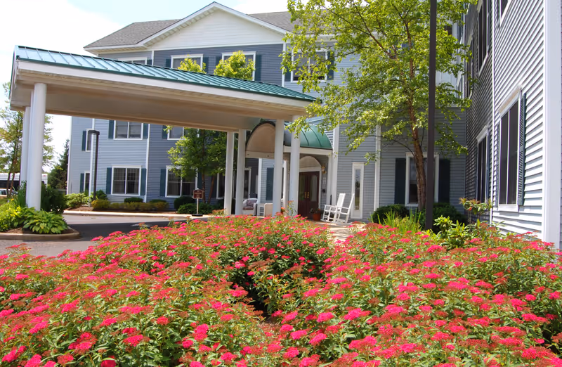 Entrance of a senior living facility with a covered driveway, surrounded by vibrant pink flowers and green shrubs. The building has light blue siding with white trim and several windows. There are two white rocking chairs near the entrance and a tree providing some shade.