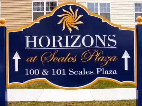 A large blue and gold sign for Horizons at Scales Plaza, located at 100 & 101 Scales Plaza, positioned outdoors on a grassy area with a building visible in the background.