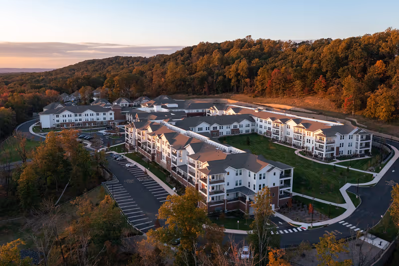 Aerial view of The Delaney of Bridgewater senior living facility surrounded by trees with autumn foliage, showing multiple connected white buildings with gray roofs, parking lots, and landscaped green areas during sunset.