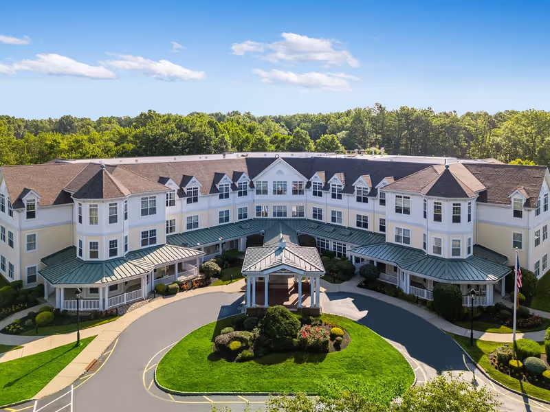A large, three-story senior living facility building with white and light yellow exterior walls, green metal awnings, and multiple windows. The building is U-shaped with a covered entrance in the center, surrounded by well-maintained landscaping and a circular driveway. Trees and a clear blue sky are visible in the background.