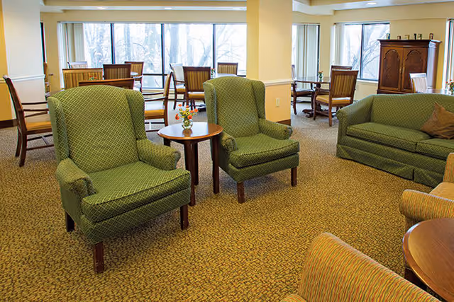 A cozy senior living facility common area with green upholstered armchairs and a matching sofa arranged around small wooden tables. In the background, there are dining tables and chairs near large windows letting in natural light.