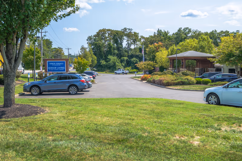Front entrance and parking lot of Arbor Terrace Middletown senior living facility with sign, cars, and landscaped grounds.
