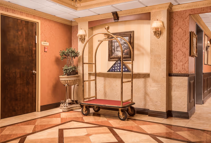 An elegant hallway corner in a senior living facility with a polished luggage cart on wheels, a potted plant on a decorative pedestal, a framed painting, and a folded American flag displayed on a shelf. The walls are adorned with patterned wallpaper and sconces with lampshades, and the floor features a geometric tile design.