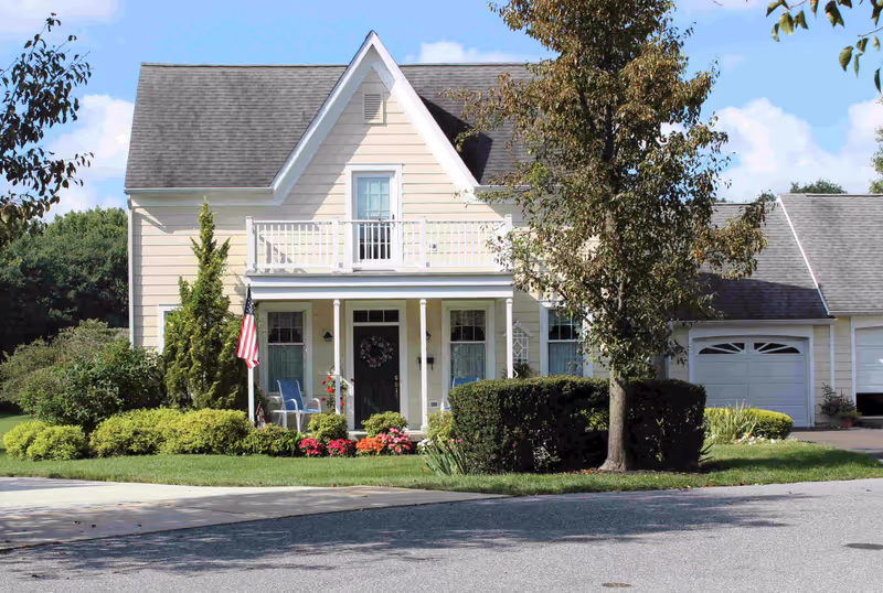Front exterior view of a two-story beige house with a small porch, white railing, American flag, and well-maintained garden with bushes and flowers. A tree is visible on the right side of the house, and there is a driveway leading to a garage.