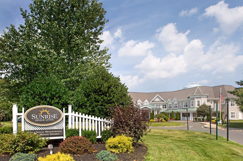 Landscaped entrance with a 'Sunrise Senior Living' sign, white fence, and the facility building in the background under a blue sky.