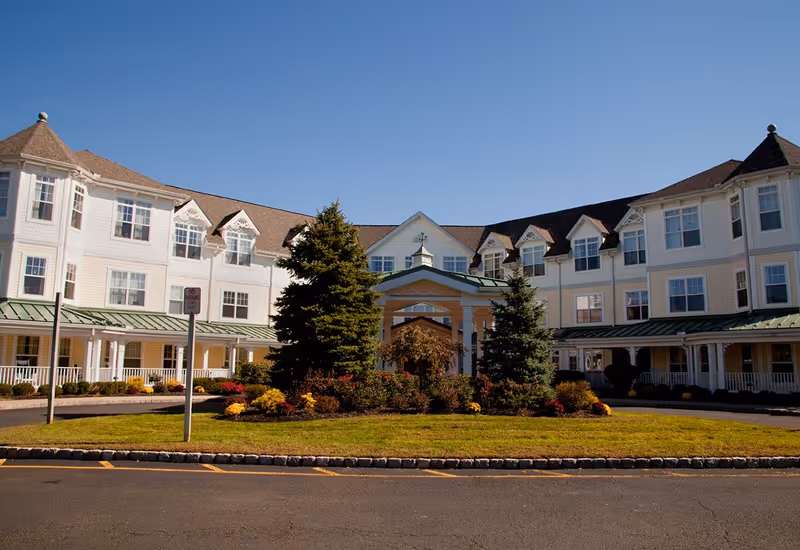 Front view of a large white multi-story senior living building with a covered entrance and landscaped circular driveway under a clear blue sky.