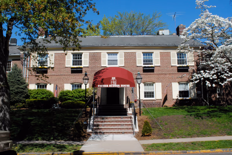 Two-story brick building with a red entrance awning labeled 'Father Hudson House', front steps, and surrounding trees and lawn.