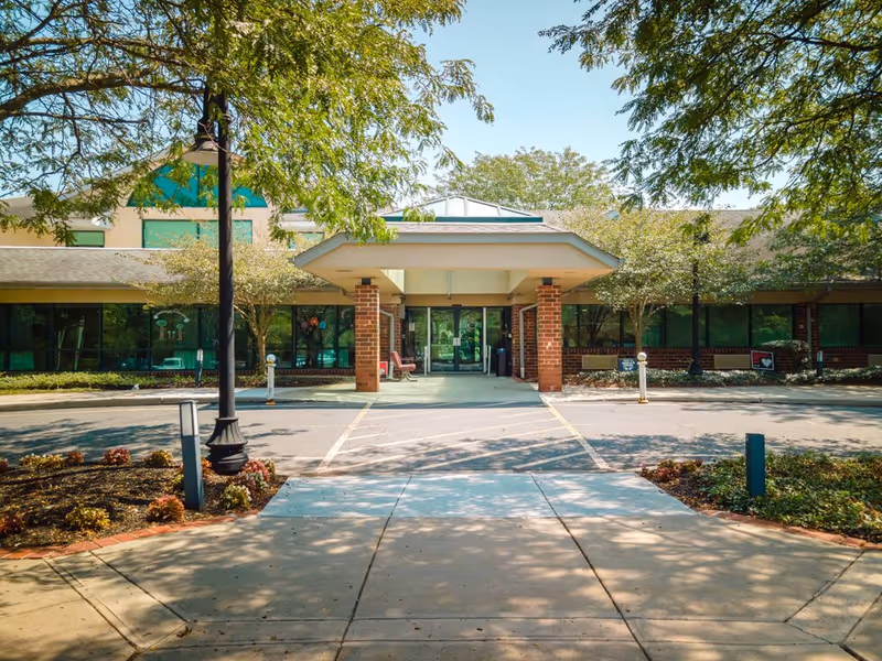 Front entrance of a senior living facility with a covered drop-off area supported by brick pillars, surrounded by trees and landscaped bushes under a clear sky.