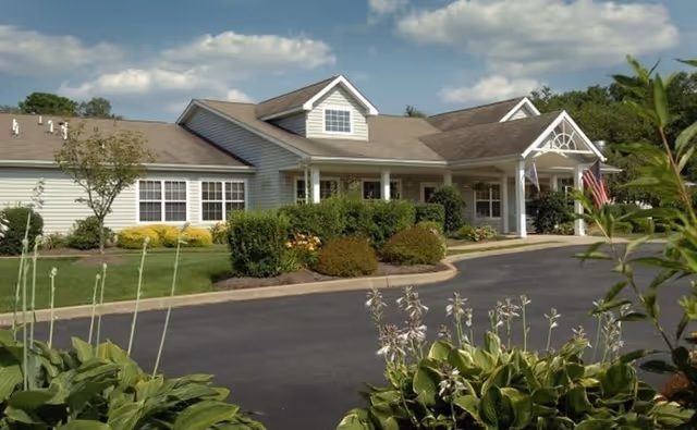Exterior view of a single-story building with a gabled roof, white siding, and a covered entrance supported by columns. The building is surrounded by well-maintained landscaping including bushes, plants, and a paved driveway. The sky is partly cloudy.