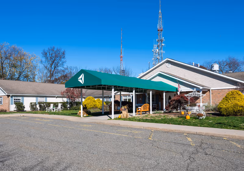 Front entrance of a single-story memory care facility with a green covered driveway, benches, landscaping and an American flag.