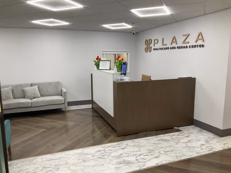 Reception area of Plaza Healthcare and Rehab Center with a front desk, sofa, flowers, and a wall sign reading PLAZA.