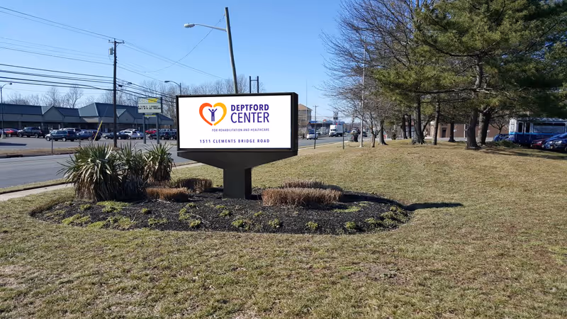 A freestanding Deptford Center sign on a grassy median by a roadside with trees and parked cars in the background.
