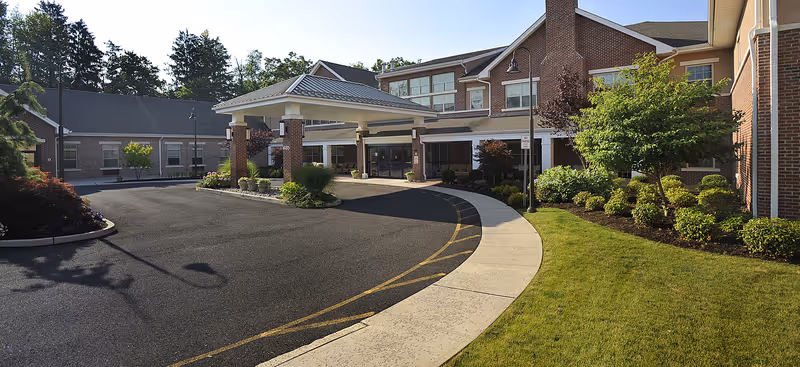 Front exterior view of a senior living facility with a covered entrance, brick buildings, landscaped greenery, and a curved driveway.