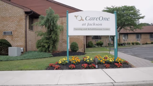 Entrance sign reading 'CareOne at Jackson Nursing and Rehabilitation Center' in front of a brick building with a flowerbed and parking area.