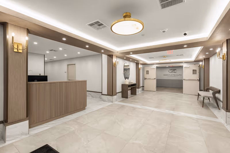 A spacious, modern reception area with a wooden front desk on the left, beige tiled floors, and soft lighting from ceiling fixtures and wall sconces. There is a bench on the right side and a console table with a round mirror and decorative items against the far wall. Double doors at the back display the Complete Care logo and name.