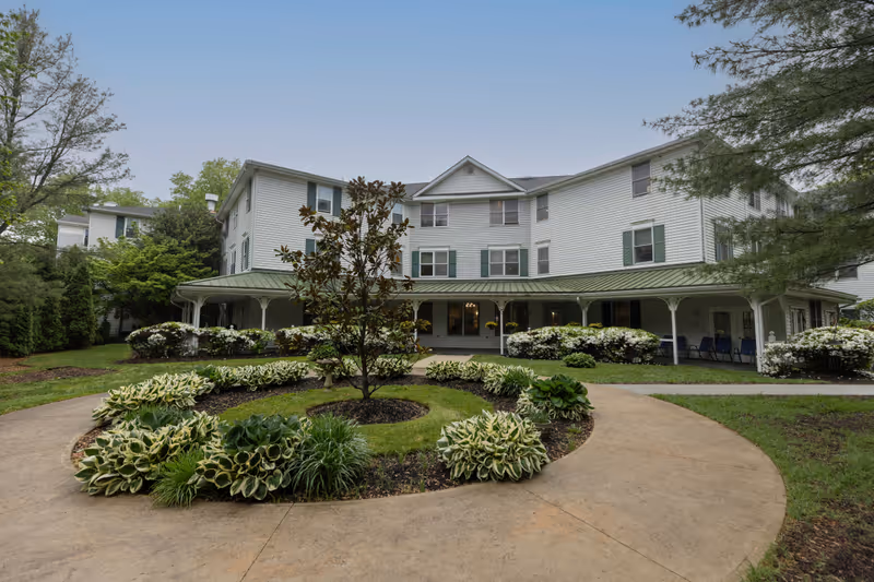 Exterior view of a three-story white building with green shutters and a green metal roof, surrounded by landscaped gardens and a circular walkway with plants and shrubs in front.