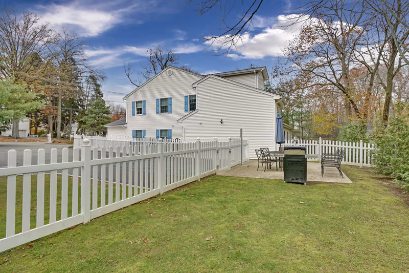 Outdoor view of a white two-story building with blue window shutters, surrounded by a white picket fence. There is a small patio area with metal chairs, a table, an umbrella, and a grill. Trees with autumn foliage are visible in the background under a partly cloudy sky.