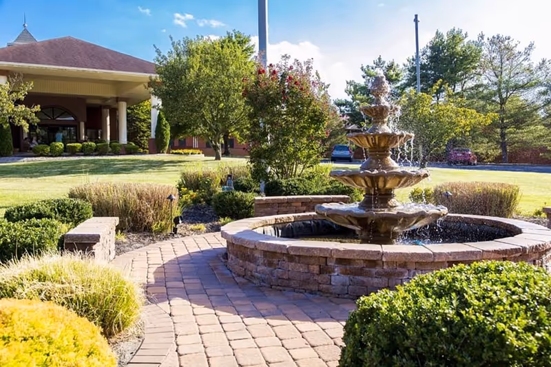 A tiered stone fountain in a landscaped brick courtyard in front of a senior living building entrance.