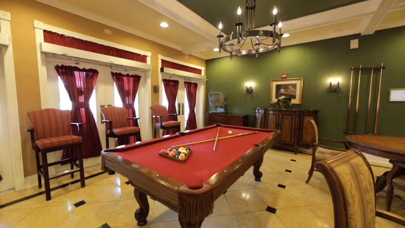A well-lit game room with a red felt pool table in the center, pool cues resting on the table, and a rack of billiard balls. The room features green walls, a chandelier overhead, several high-backed chairs with red and gold upholstery, and windows with red curtains allowing natural light to enter. There is also a wooden cabinet and a small TV in the corner.