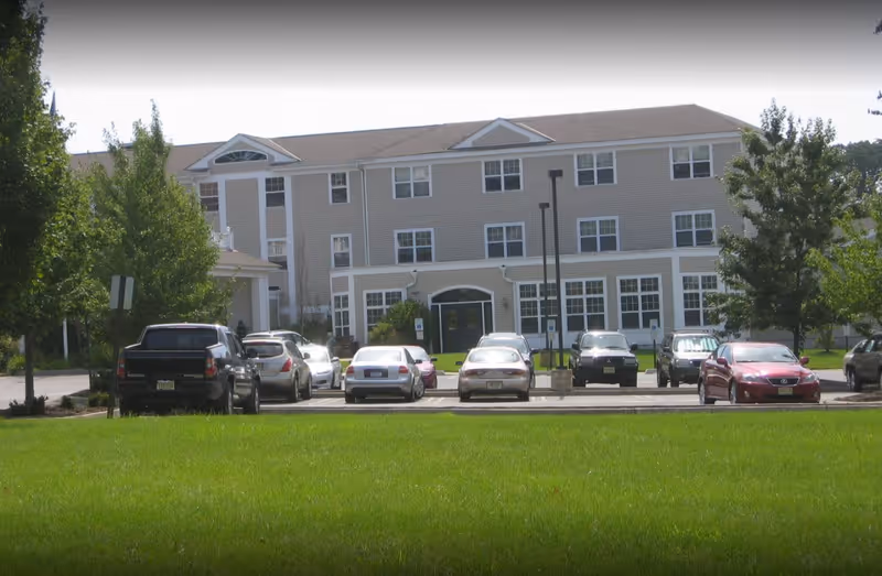 Front exterior view of a three-story assisted living facility building with multiple windows, a parking lot with several cars, green grass lawn, and trees on either side.