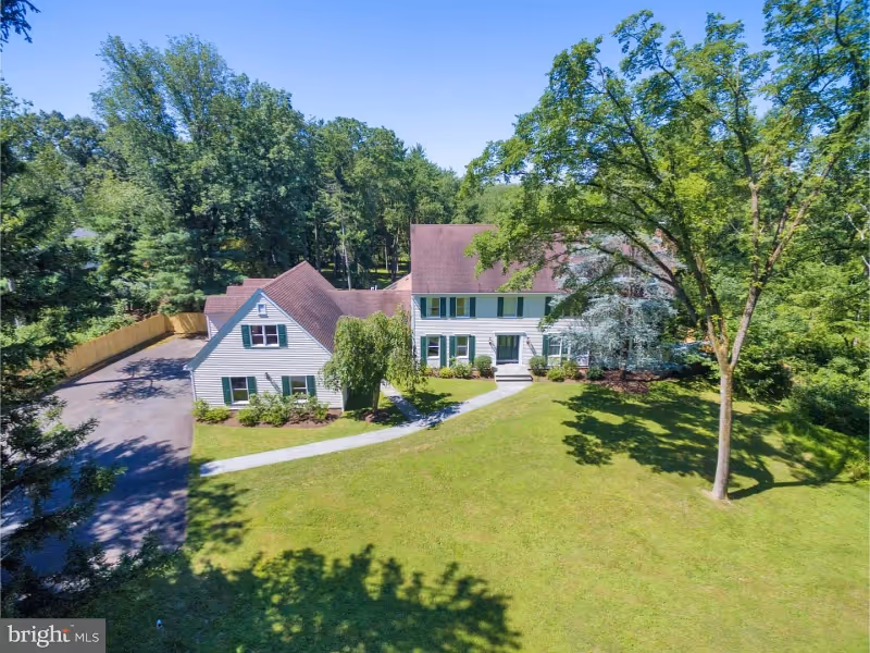 A large white two-story house with green shutters and a brown roof, surrounded by a well-maintained lawn and tall trees under a clear blue sky. A paved driveway leads to the house, and a sidewalk curves from the driveway to the front door.