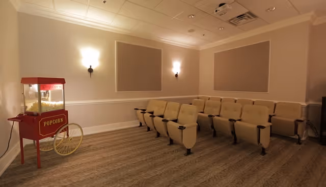 A small theater room with two rows of beige cushioned seats with cup holders, a popcorn machine on the left side, beige walls with two wall sconces, and a patterned carpeted floor.