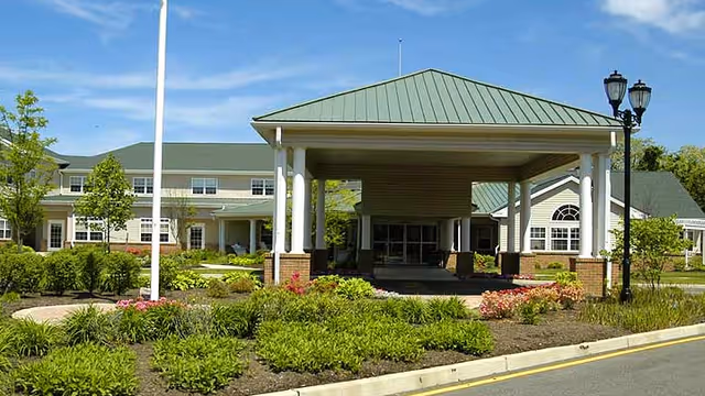 Front exterior view of The Orchards Assisted Living facility featuring a covered entrance with white columns, a green metal roof, landscaped garden beds with shrubs and flowers, and a black street lamp on the right side.