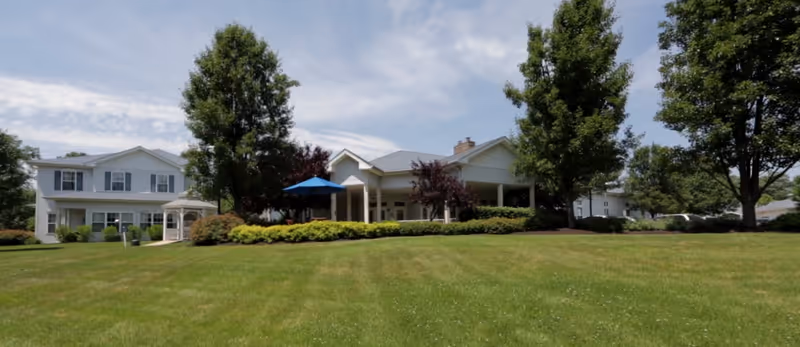 Front exterior of a two-story light-colored building with a wide lawn, trees, a gazebo, and a covered entrance with a blue umbrella.