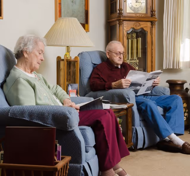 An elderly woman and an elderly man sitting in blue armchairs in a cozy living room. The woman is reading a book while the man is reading a newspaper. There is a wooden grandfather clock and a table with a lamp between them. The room has beige walls and carpet, with curtains covering a window.
