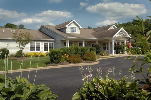 Exterior view of a single-story senior living facility building with white siding, a covered entrance, and well-maintained landscaping including bushes and flowering plants under a partly cloudy sky.