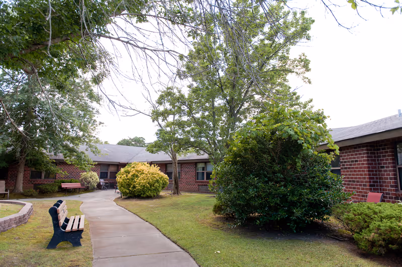 A courtyard walkway between single-story brick buildings lined with benches, trees, and shrubs.
