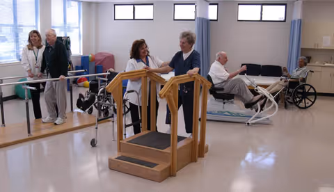 A rehabilitation room in a nursing center where elderly individuals are engaged in physical therapy activities. One elderly woman is assisted by a therapist while walking on a wooden step platform with handrails. Another elderly man is walking with support bars and a therapist nearby. In the background, two elderly men are seated, one in a wheelchair and the other on an exercise device. The room is well-lit with large windows and has medical and therapy equipment.