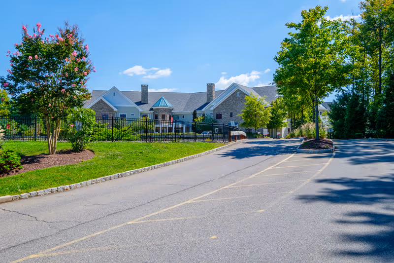 View of a senior living facility building named Parker at Monroe, with a paved driveway leading up to it, surrounded by green trees and landscaping under a blue sky with some clouds.