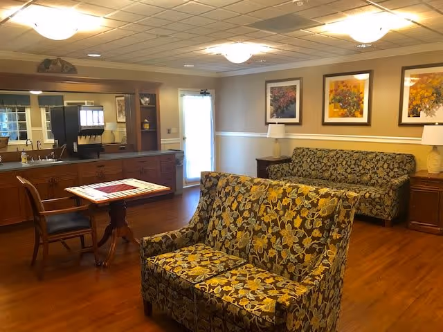 A cozy senior living common area with floral patterned sofas, a wooden table with chairs, a countertop with a coffee machine, and framed floral artwork on the walls. The room has wooden flooring and soft ceiling lights.