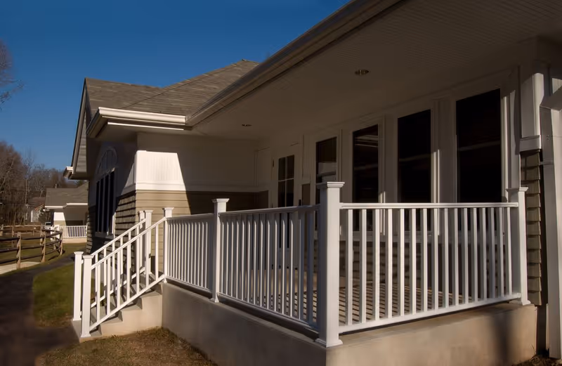 Front porch of a single-story building with white railing, steps, and windows under a covered entrance.