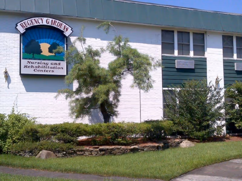 Exterior view of a building with white brick walls and green paneling. There is a sign on the building that reads 'Regency Gardens Nursing and Rehabilitation Centers'. The foreground features a landscaped area with bushes, a small tree, rocks, and a grassy lawn.