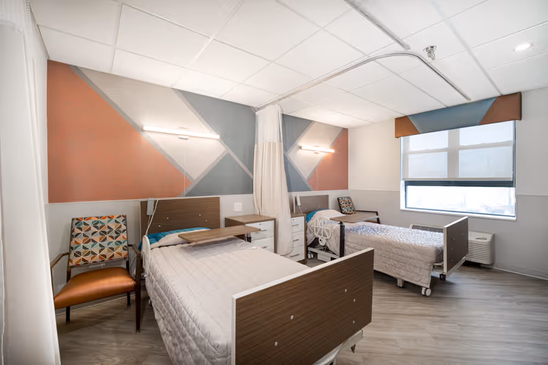 A bright and clean healthcare facility room with two hospital beds separated by a privacy curtain. Each bed has a wooden headboard and footboard, beige quilted bedding, and an overbed table. There are two patterned chairs next to each bed, a window with a roller shade and valance, and modern geometric wall art in shades of orange, gray, and blue. The floor is light wood, and the ceiling has white tiles with fluorescent lighting.