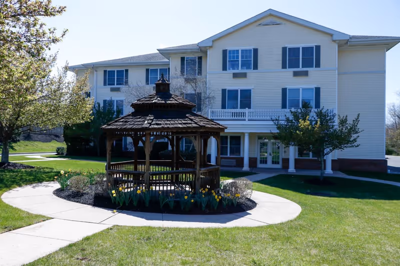 A three-story light yellow building with multiple windows and a white entrance door, surrounded by green grass and trees. In front of the building, there is a wooden gazebo with a shingled roof, encircled by a curved concrete walkway and flower beds with yellow flowers.