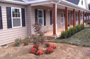 Exterior view of a single-story building with beige siding, white trim, and a covered porch supported by wooden columns. There is a small landscaped area in front with a young tree surrounded by red flowers and some small green shrubs near the building.