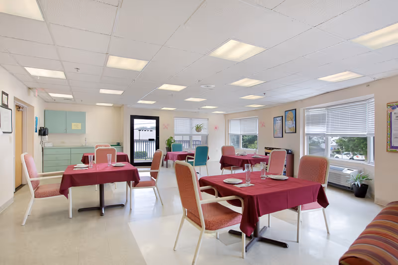 A bright dining room with several tables covered in burgundy tablecloths, each set with plates, glasses, and utensils. The room has large windows with blinds, allowing natural light to enter. There are pink cushioned chairs around the tables, a small kitchenette area with green cabinets, and some plants and framed pictures on the walls.