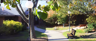 Outdoor garden area at Autumn Lake Healthcare at Oceanview featuring a paved walkway, trees, bushes, lamp posts, and wooden benches with a building partially visible in the background.