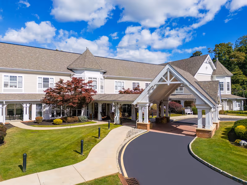 Front exterior view of a senior living facility named Sunrise of Lincroft, featuring a large covered entrance with white pillars, well-maintained green lawns, shrubs, and trees under a partly cloudy blue sky.